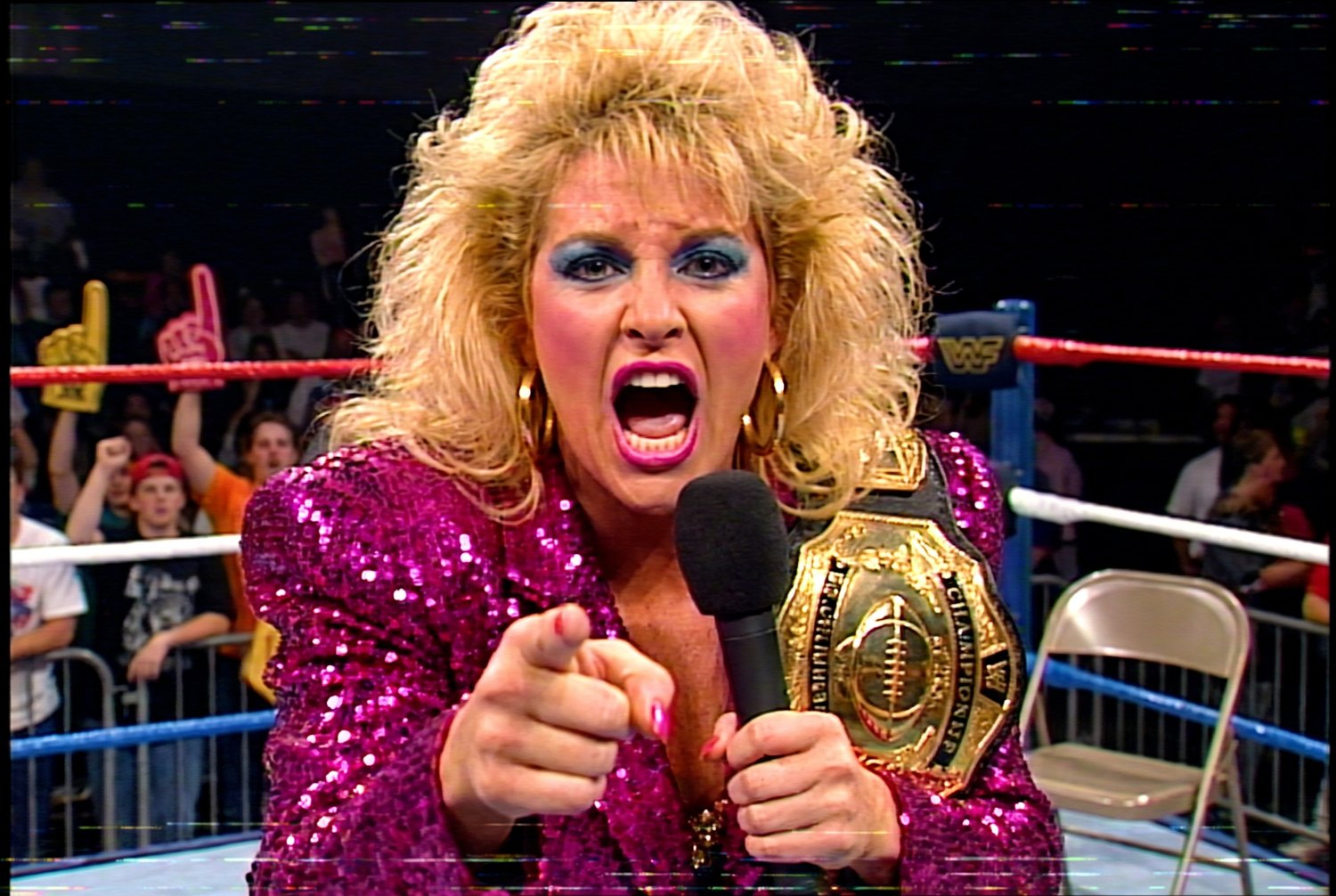 Vixen Vance at ringside in a sequined magenta jacket, screaming into a microphone and pointing at the camera, championship belt with a football on the plate over her shoulder
