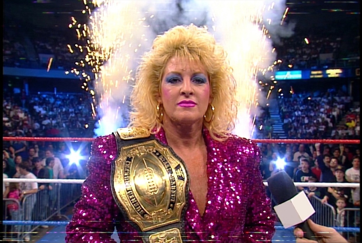 Vixen Vance in the centre of a wrestling ring with a gold championship belt with a football on the plate over her shoulder, gold pyro erupting behind her