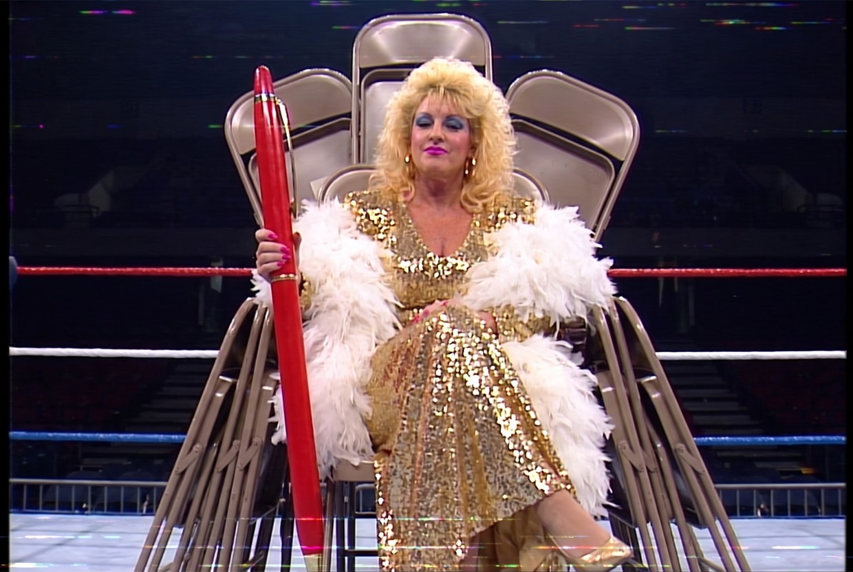Vixen Vance in a gold sequined gown and white feather boa seated on a throne built of stacked folding chairs in an empty arena