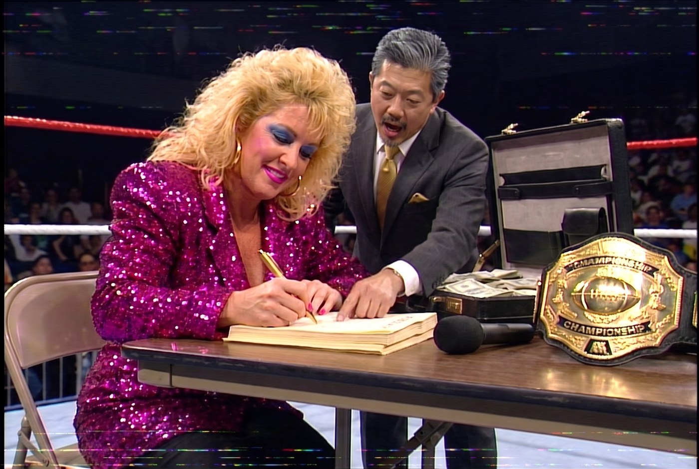 Vixen Vance at a ringside table signing a consulting contract, an open briefcase of cash and the championship belt beside her
