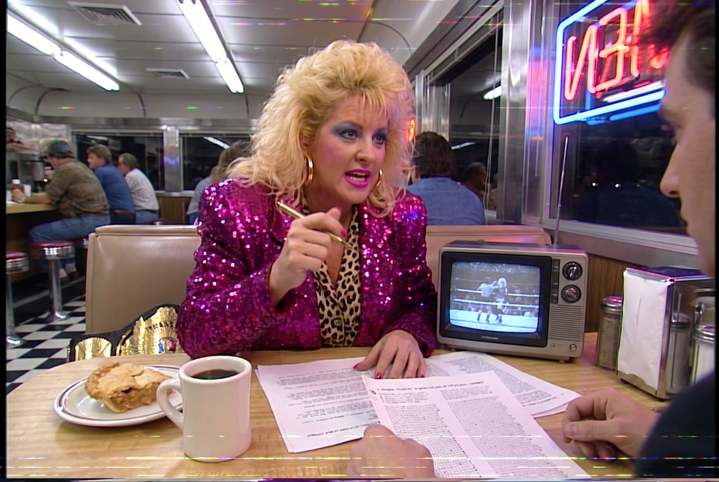 Vixen Vance working out of a diner booth covered in printouts, a portable TV showing the draft, pen in hand