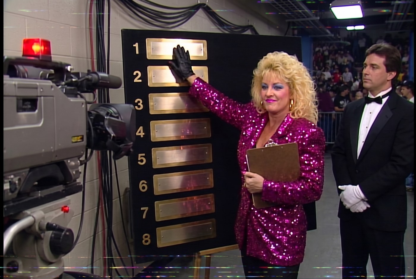 1987 broadcast still: Vixen Vance backstage beside a numbered board of eight brass nameplates, hand on slot one, a television camera rolling on her