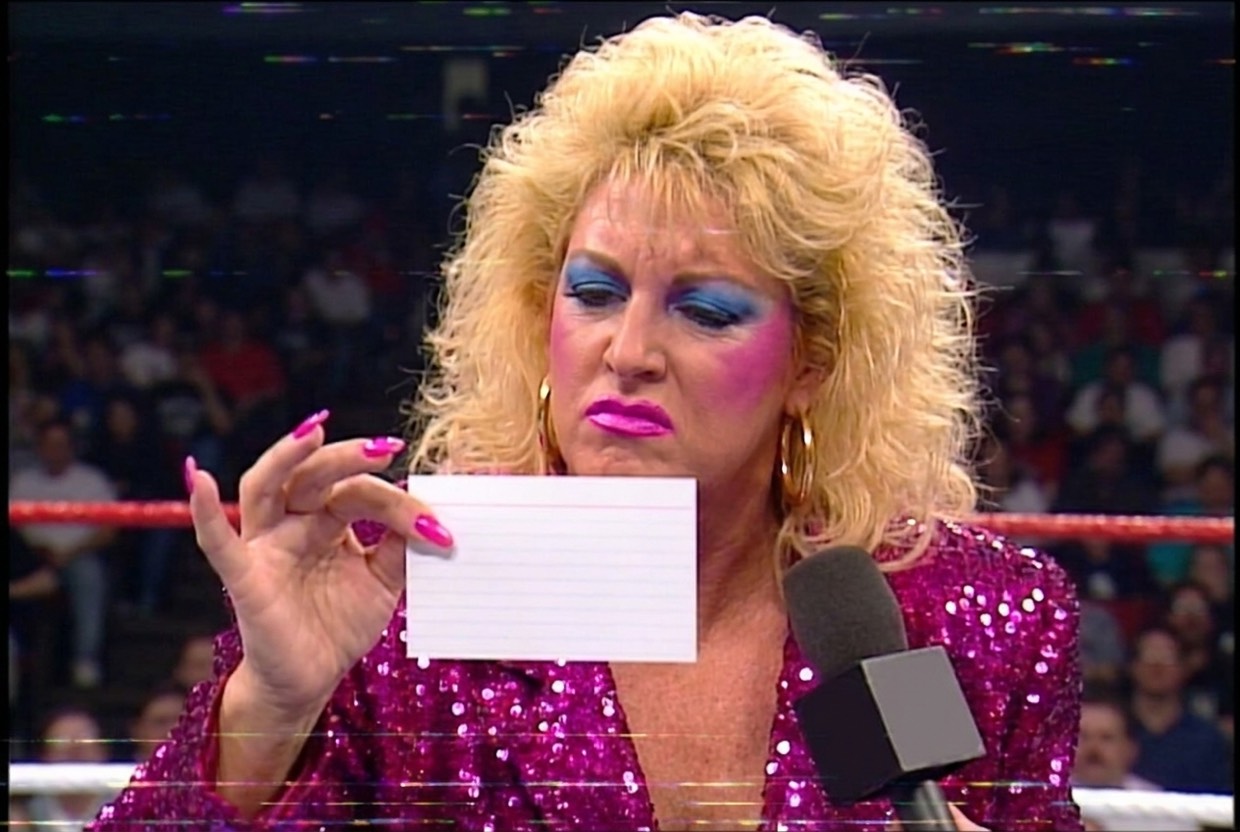 Vixen Vance at ringside holding a small blank index card at arm's length between two fingernails, squinting at it with contempt