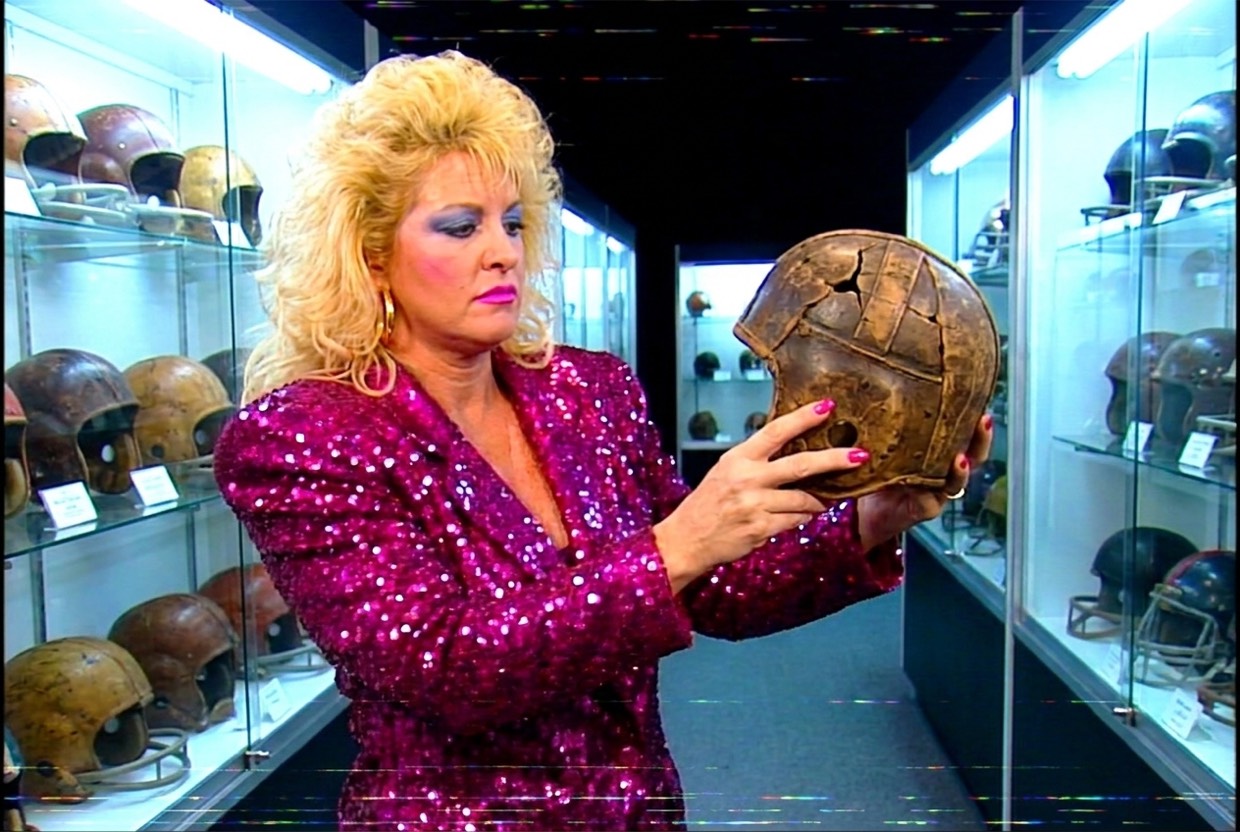 Vixen Vance in a trophy hall of antique leather football helmets, holding a cracked ancient helmet at arm's length with an expression of pity