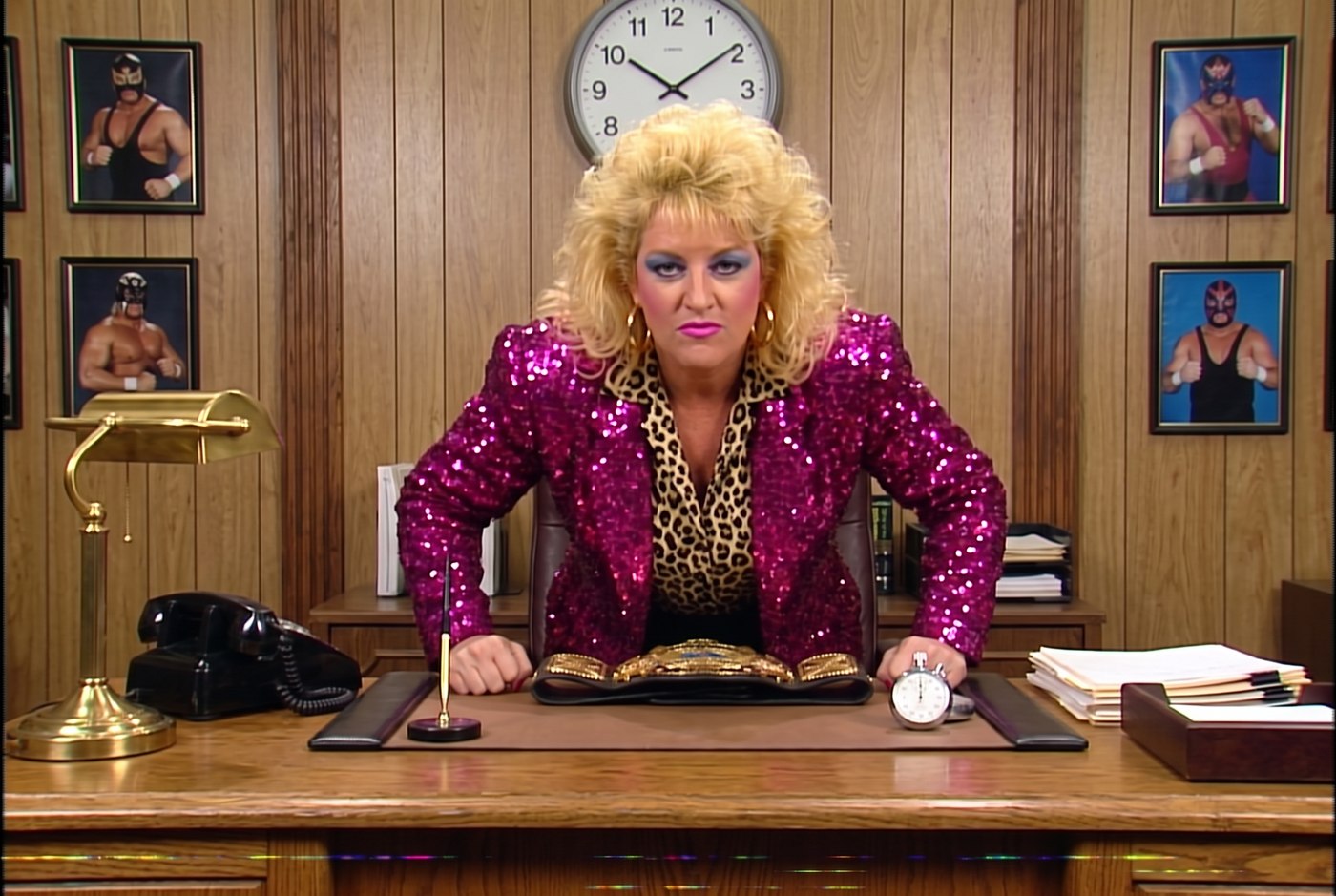 Vixen Vance in the front office in front of a wall of two dozen clocks, holding up a stopwatch