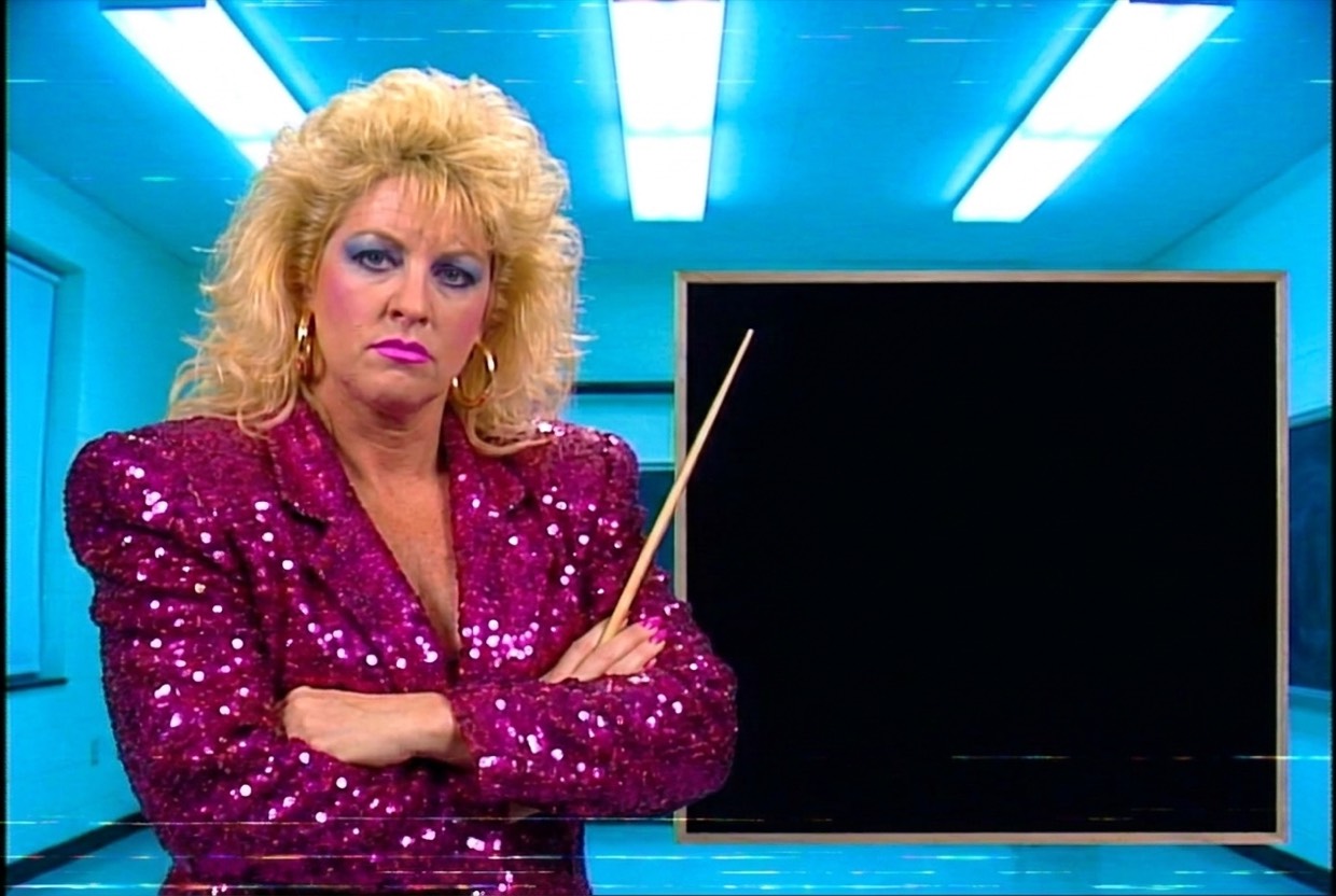 Vixen Vance beside a blank chalkboard with arms folded and a wooden pointer, wearing a stern schoolteacher expression